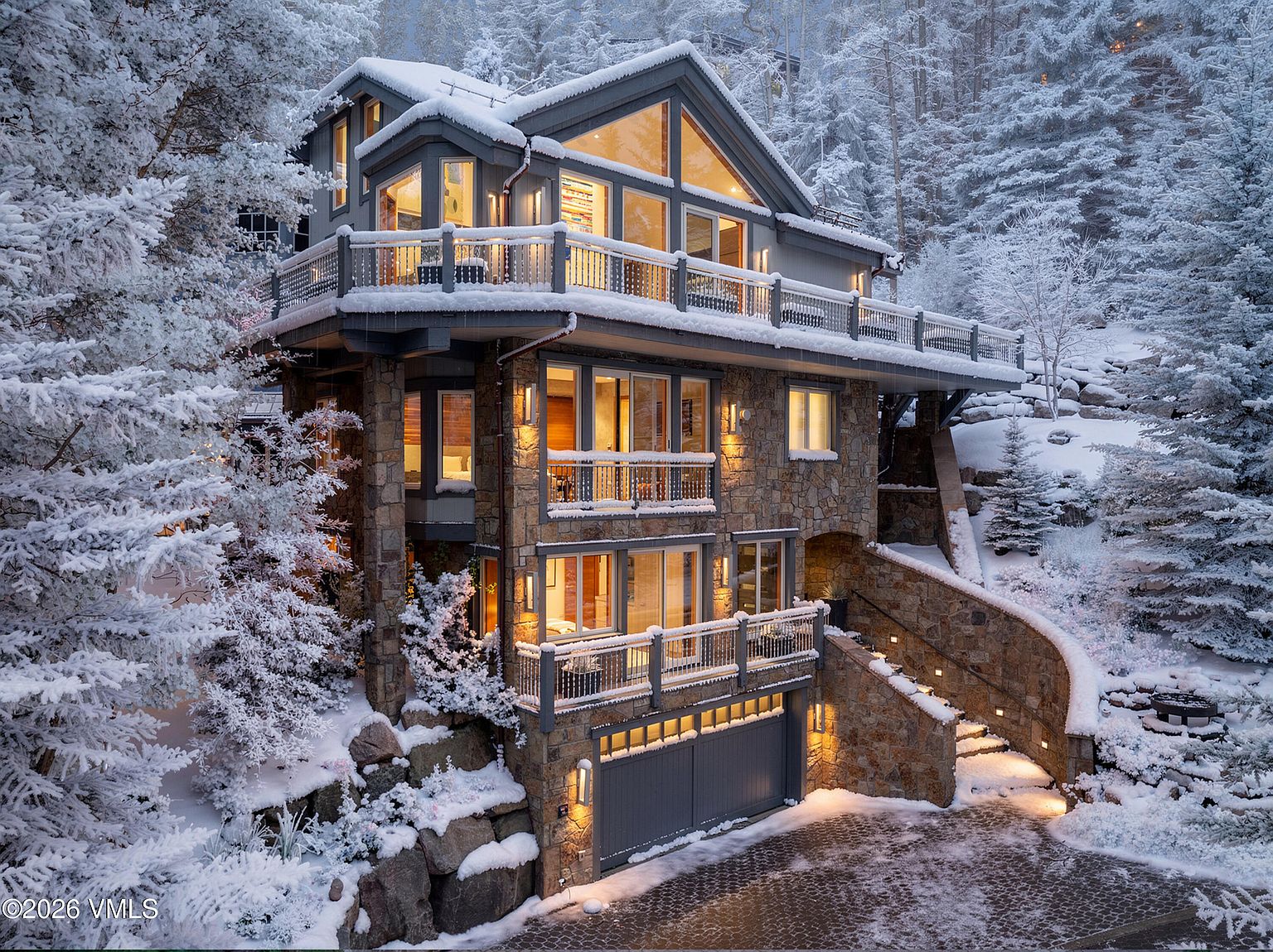 This is a stunning front view of a multi-story luxury home nestled in a snowy, wooded landscape. The house features a stone facade, multiple balconies with wooden railings, and large windows that offer ample natural light. The driveway and surrounding landscape are covered in snow, creating a picturesque winter scene.