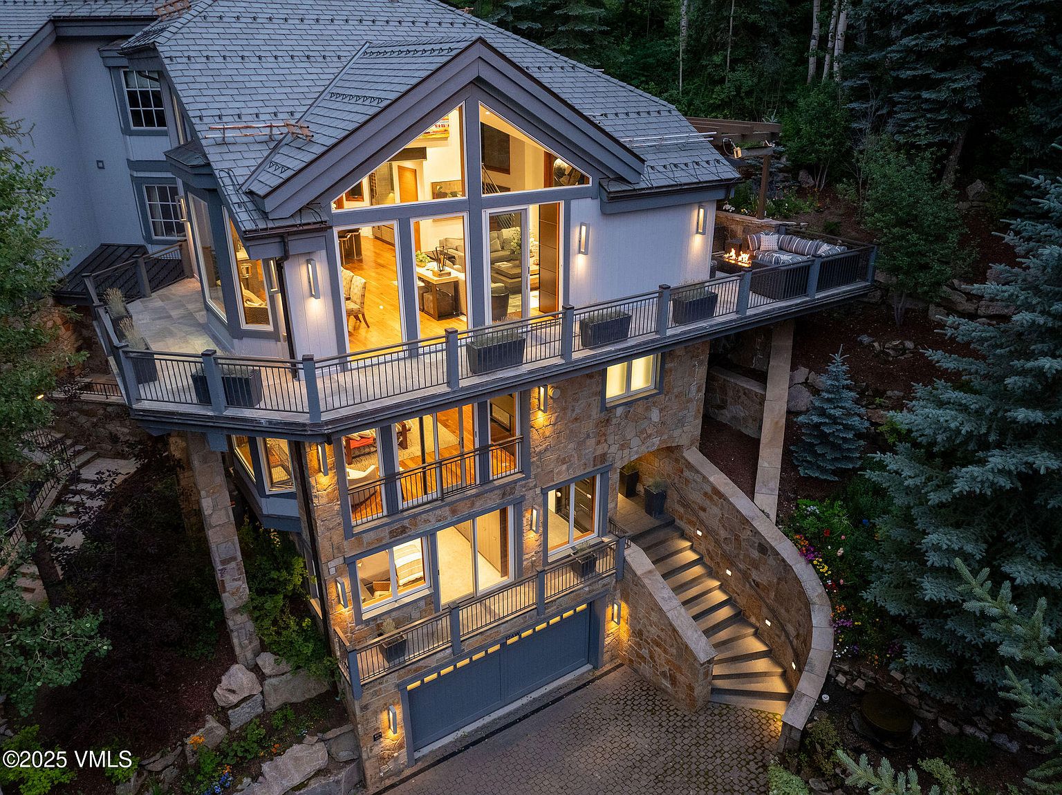 This is an aerial view of a luxurious multi-story home with a stone and stucco exterior. The house features multiple balconies with metal railings, large windows offering views of the interior, and a well-manicured landscape with stone steps leading to the entrance. The overall impression is one of sophisticated elegance and architectural grandeur.