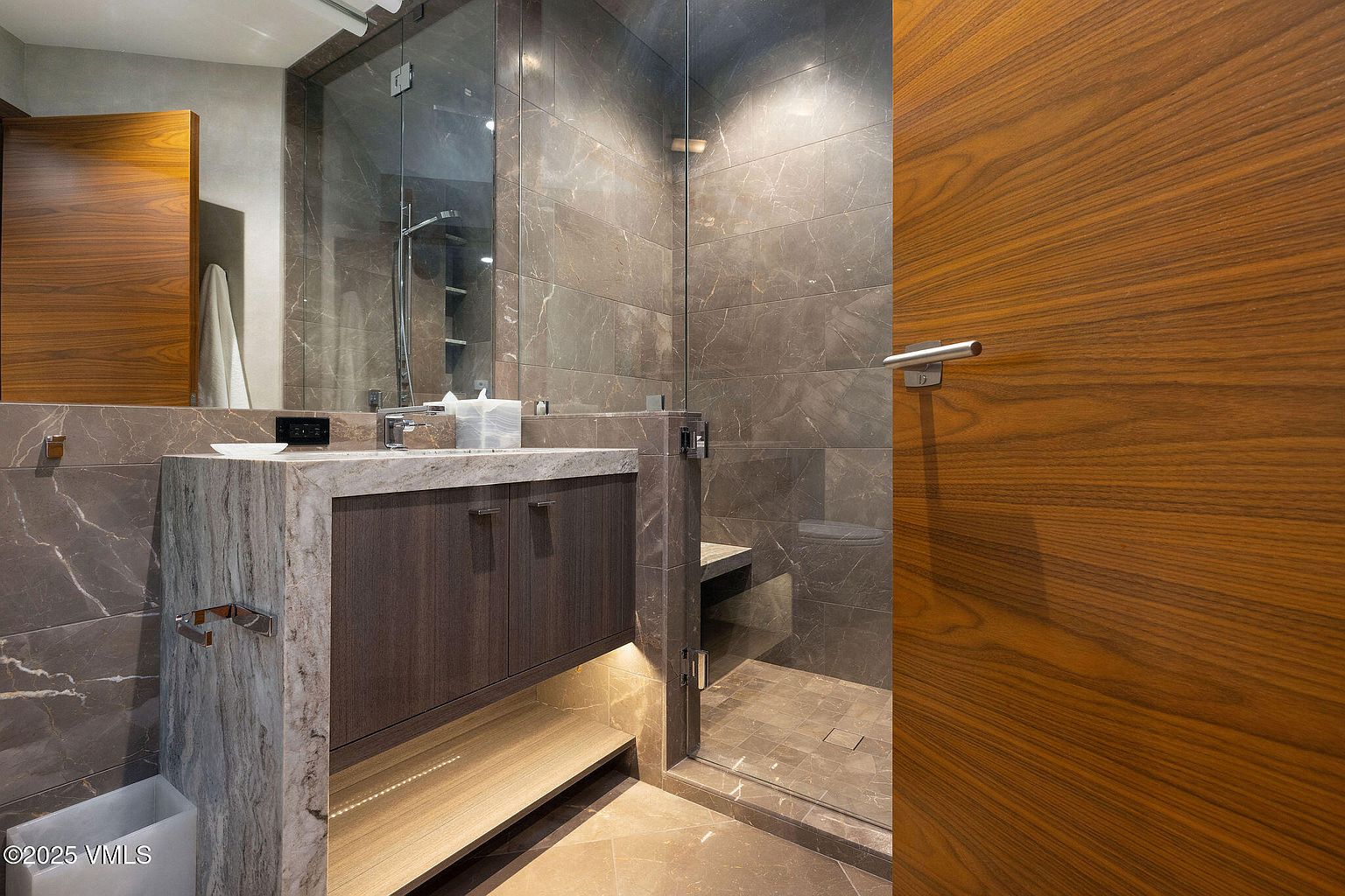 This is a primary bathroom featuring a modern design with marble-like tile on the walls and floor. The vanity has a stone countertop and dark wood cabinets, with a shelf underneath illuminated by soft lighting. A glass-enclosed shower is visible, and a wooden door with a silver handle is on the right side of the frame.
