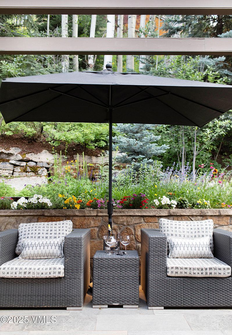 This image showcases a cozy outdoor patio setting, featuring two wicker chairs with patterned cushions flanking a small matching table. A large black umbrella provides shade, while a stone retaining wall with colorful flowers and lush greenery creates a serene backdrop. The scene evokes a sense of relaxation and outdoor living.