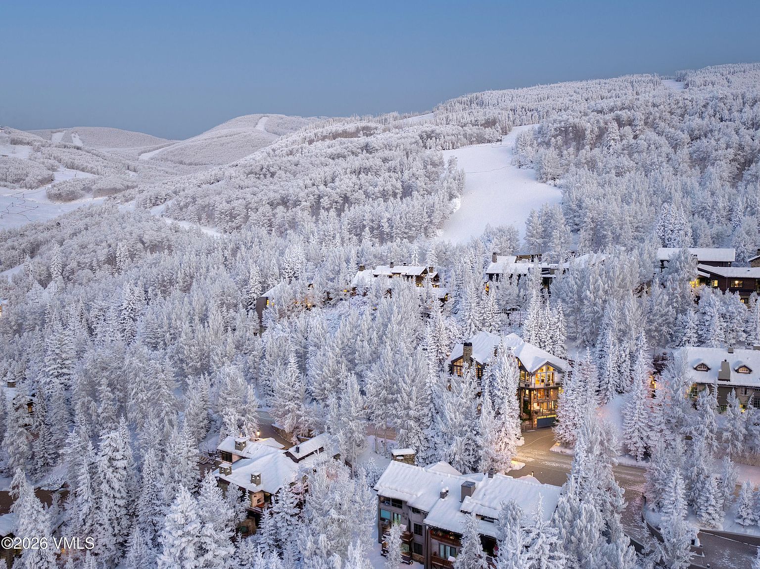 This aerial view showcases a luxurious mountain resort covered in fresh snow. Several chalet-style homes with warmly lit windows are nestled among snow-laden evergreen trees. The scene evokes a sense of winter tranquility and upscale living, perfect for attracting buyers seeking a serene retreat.