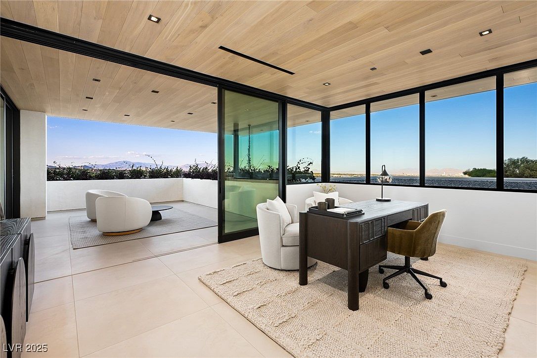 This is a modern office space featuring a dark wood desk, a comfortable chair, and a large window offering a scenic view. The room has a light wood ceiling and a neutral-toned rug, creating a bright and inviting atmosphere. Sliding glass doors open to an outdoor seating area, seamlessly blending indoor and outdoor spaces.