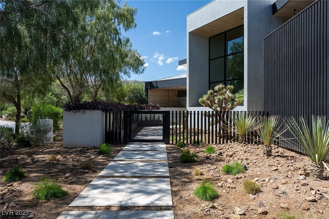 This image showcases the entryway of a modern home, featuring a pathway constructed of large, square concrete pavers leading to a black metal gate. The landscaping includes drought-resistant plants and trees, complementing the contemporary architectural style of the house with its clean lines and minimalist design. The exterior of the home is a combination of gray stucco and dark vertical siding, creating a sophisticated and inviting entrance.