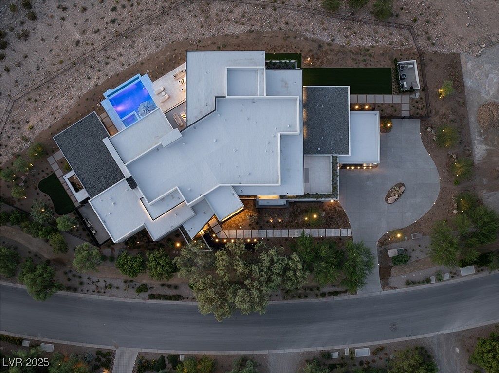 This aerial view showcases a modern luxury home with a unique architectural design, featuring multiple flat roof sections, a swimming pool with a spa, and meticulously landscaped grounds. The property includes a spacious driveway and is surrounded by desert landscaping, offering privacy and a sense of exclusivity. The lighting highlights the clean lines and sophisticated style of the residence.