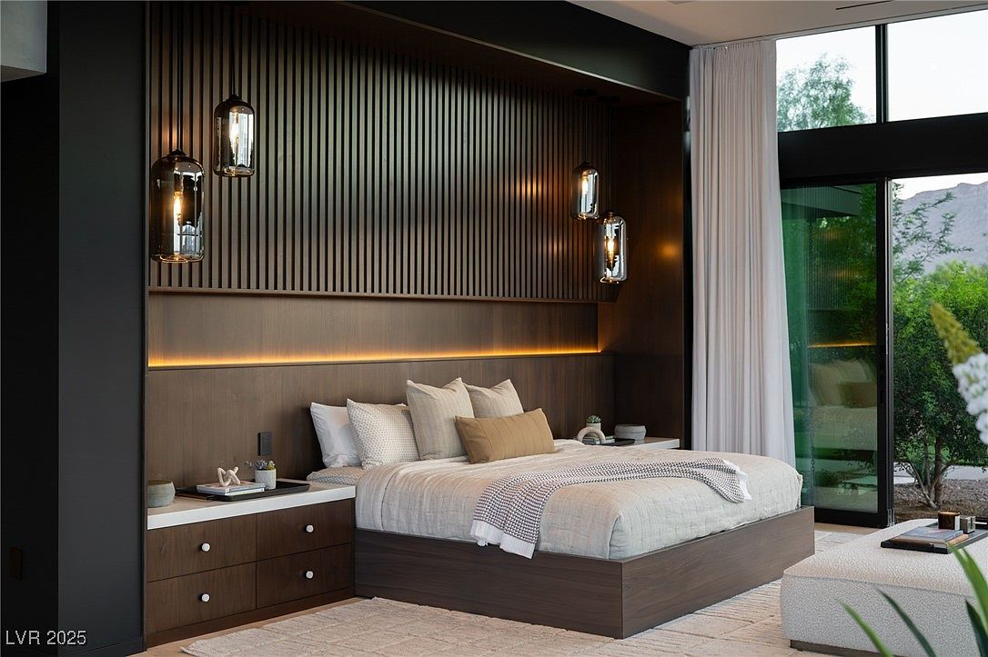 This is a beautifully designed primary bedroom featuring a modern aesthetic. The room showcases a dark wood paneled headboard with integrated lighting, complemented by stylish pendant lights. A large bed with neutral bedding and a cozy throw sits atop a light-colored rug, creating a serene and inviting atmosphere, with a view to the outside.
