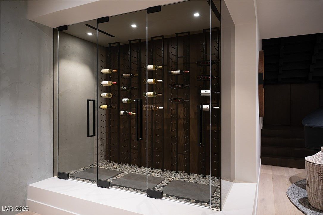 This image showcases a modern wine cellar enclosed by glass panels, featuring a concrete accent wall and a dark wood wine rack system. The floor is covered with small pebbles and stepping stones, adding a unique textural element. The cellar is well-lit with recessed lighting, creating an elegant and sophisticated space for wine storage.