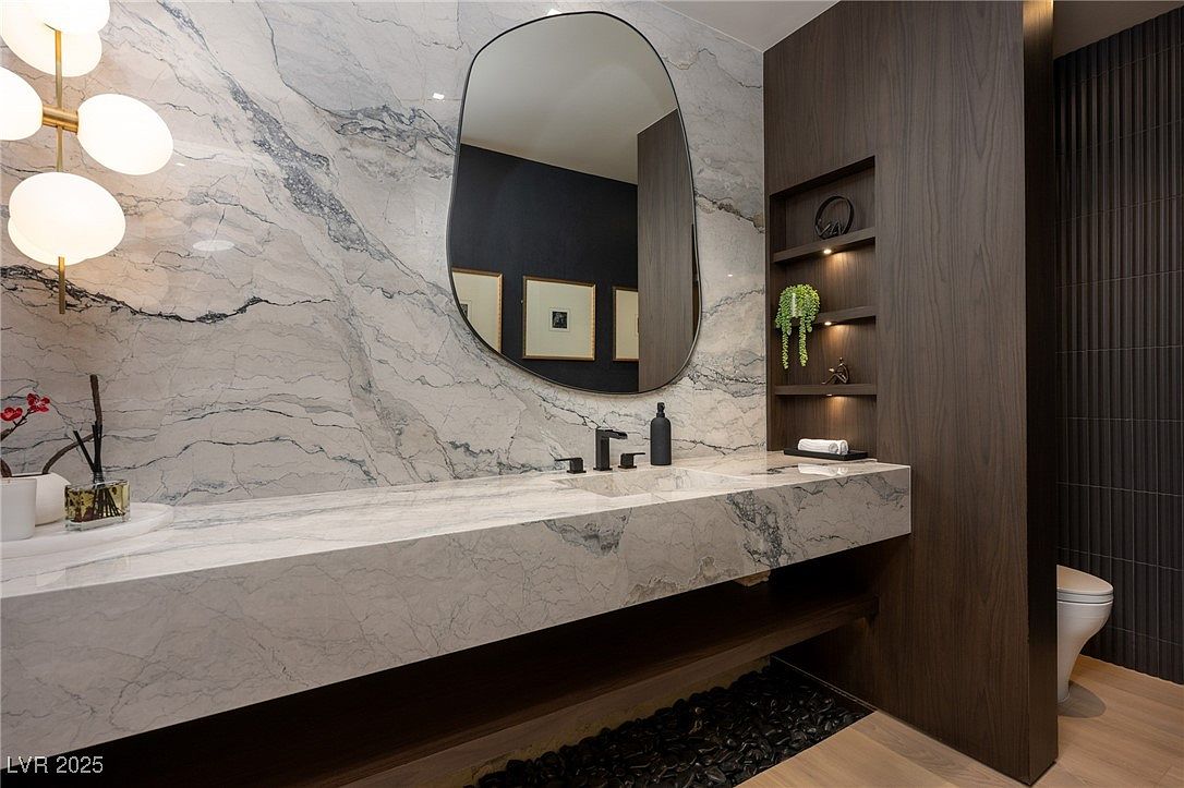 This is a modern bathroom featuring a marble countertop and backsplash with a unique, organically shaped mirror. Dark wood cabinetry with built-in shelving adds a touch of sophistication, while the black pebble detail beneath the floating vanity creates visual interest. The overall design is sleek and luxurious.