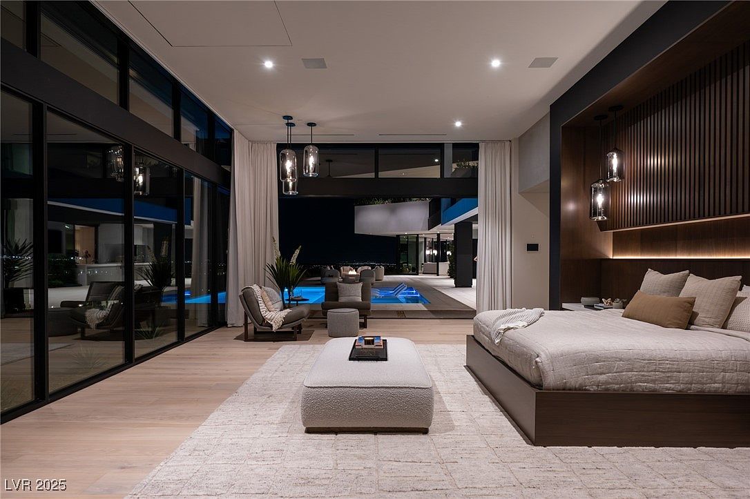 This is a luxurious primary bedroom with a modern design that seamlessly blends indoor and outdoor living. The room features floor-to-ceiling windows offering views of a private pool and outdoor lounge area. The interior is elegantly furnished with a large bed, a comfortable seating area, and stylish lighting fixtures, creating a serene and sophisticated atmosphere.