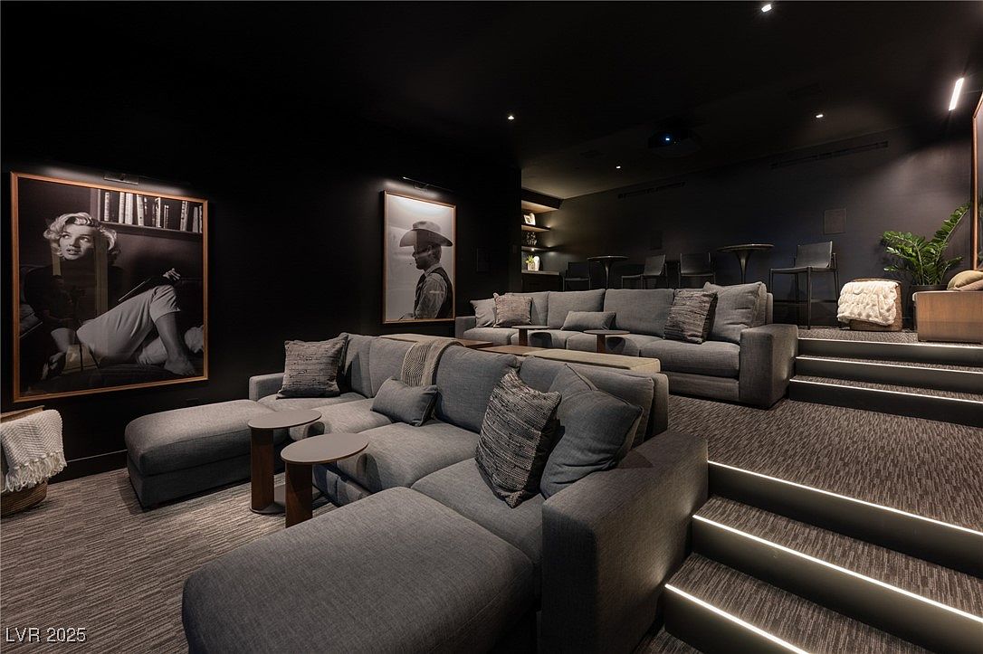 This is a luxurious home theater room featuring tiered seating with plush gray sofas and accent pillows. The walls are adorned with framed black and white portraits, and the room is illuminated by subtle recessed lighting and integrated step lighting. The overall impression is one of comfort and sophistication, perfect for a high-end cinematic experience.