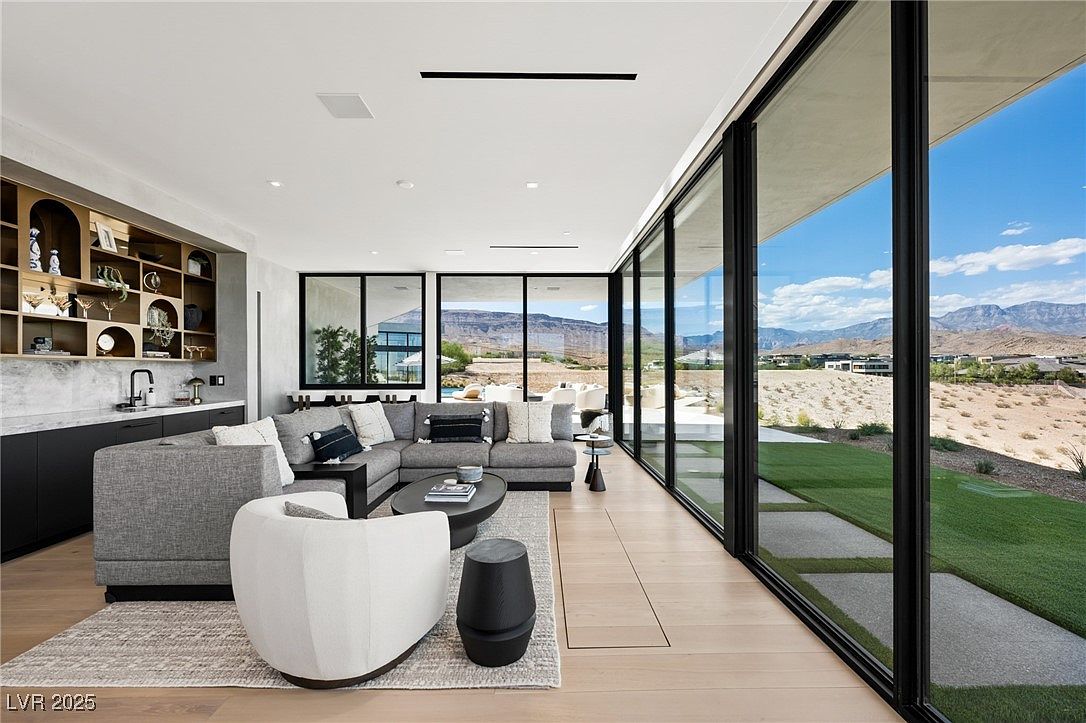 This is a bright and modern living room featuring a large sectional sofa, a white accent chair, and a dark wood side table. Floor-to-ceiling windows offer stunning views of the landscape, bringing the outdoors in. A built-in bar area with shelving adds a touch of sophistication and functionality to the space.