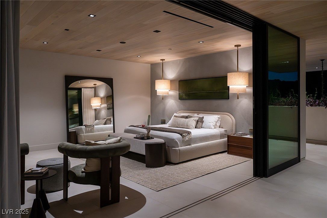 This is a luxurious primary bedroom featuring a neutral color palette, a large bed with multiple pillows, and modern lighting fixtures. The room has a minimalist aesthetic with a large mirror and a sliding glass door leading to an outdoor space. The overall impression is one of comfort and sophistication.