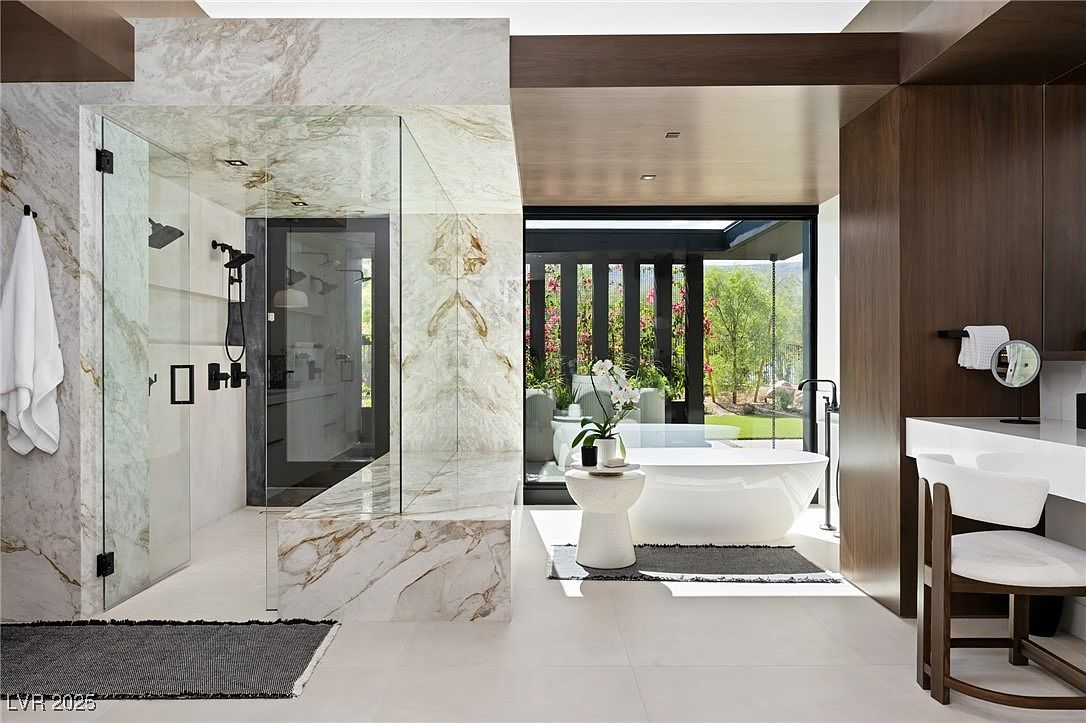 This is a luxurious primary bathroom featuring a glass-enclosed shower with marble walls and modern fixtures. A freestanding bathtub sits near a large window offering a view of the outdoor landscape. The bathroom also includes a vanity area with a chair and a dark wood accent wall, creating a spa-like atmosphere.