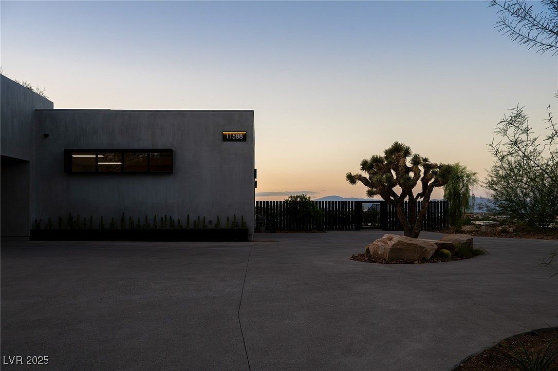 This image showcases the front exterior of a modern home at dusk. The building features a minimalist design with a flat roof, concrete walls, and a horizontal window. A Joshua tree and desert landscaping add to the property's unique appeal, while the address number is prominently displayed on the facade.