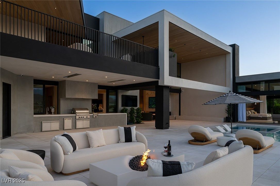 This image showcases a luxurious outdoor patio area with modern architectural design. The space features comfortable white lounge furniture, a fire pit, and an outdoor kitchen with a grill. A swimming pool is visible in the background, along with a covered balcony and sleek, contemporary building design, creating an inviting and upscale outdoor living space.