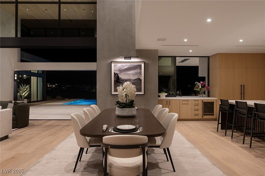 This interior shot showcases a modern dining room with a long, dark wood table surrounded by light-colored chairs with black legs. A large black and white photograph hangs on the wall above the table, and a floral arrangement serves as a centerpiece. The room opens to an outdoor area with a pool, creating a seamless indoor-outdoor living experience.