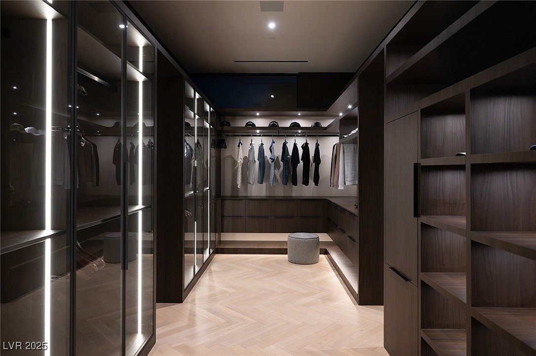 This is a well-organized walk-in closet featuring dark wood cabinetry and shelving, illuminated by vertical LED lighting strips. Clothes hang neatly on rods, and hats are displayed on an upper shelf, while a herringbone-patterned wood floor adds a touch of elegance. The closet offers ample storage space and a luxurious feel.
