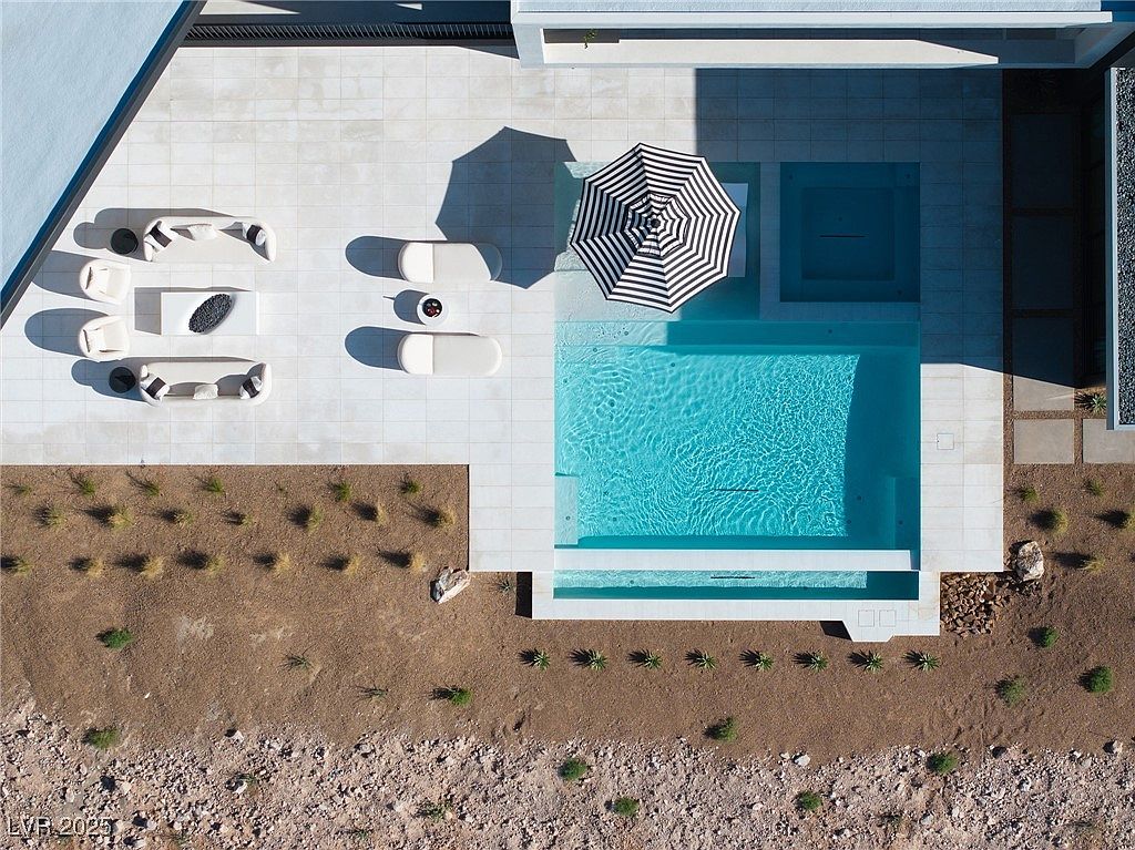 This is an aerial view of a modern backyard featuring a rectangular pool with a built-in spa. The pool area is surrounded by white tile and includes a striped umbrella, lounge chairs, and an outdoor seating area with a fire pit. The landscaping consists of desert plants and gravel, creating a clean and contemporary aesthetic.
