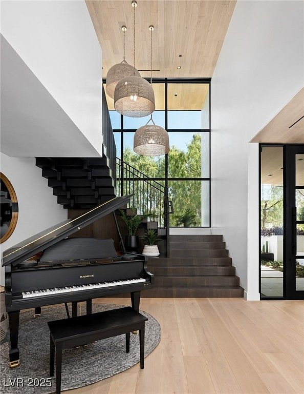 This interior shot showcases a grand entryway with a striking staircase design, featuring a black grand piano and a modern aesthetic. The space is illuminated by unique pendant lighting, complemented by large windows that bring in natural light and offer a view of the outdoors. The combination of dark and light elements creates a sophisticated and inviting atmosphere.