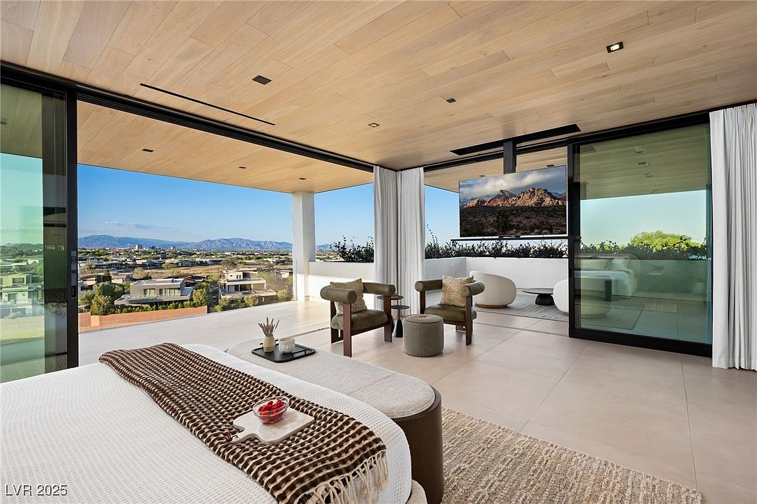 This is a luxurious primary bedroom featuring a large bed with a brown and white throw, a seating area with two chairs and a small table, and a flat-screen TV mounted on a ceiling fixture. Sliding glass doors open to a balcony with city and mountain views, creating a seamless indoor-outdoor living experience. The room is spacious and well-lit, with a modern and minimalist design.