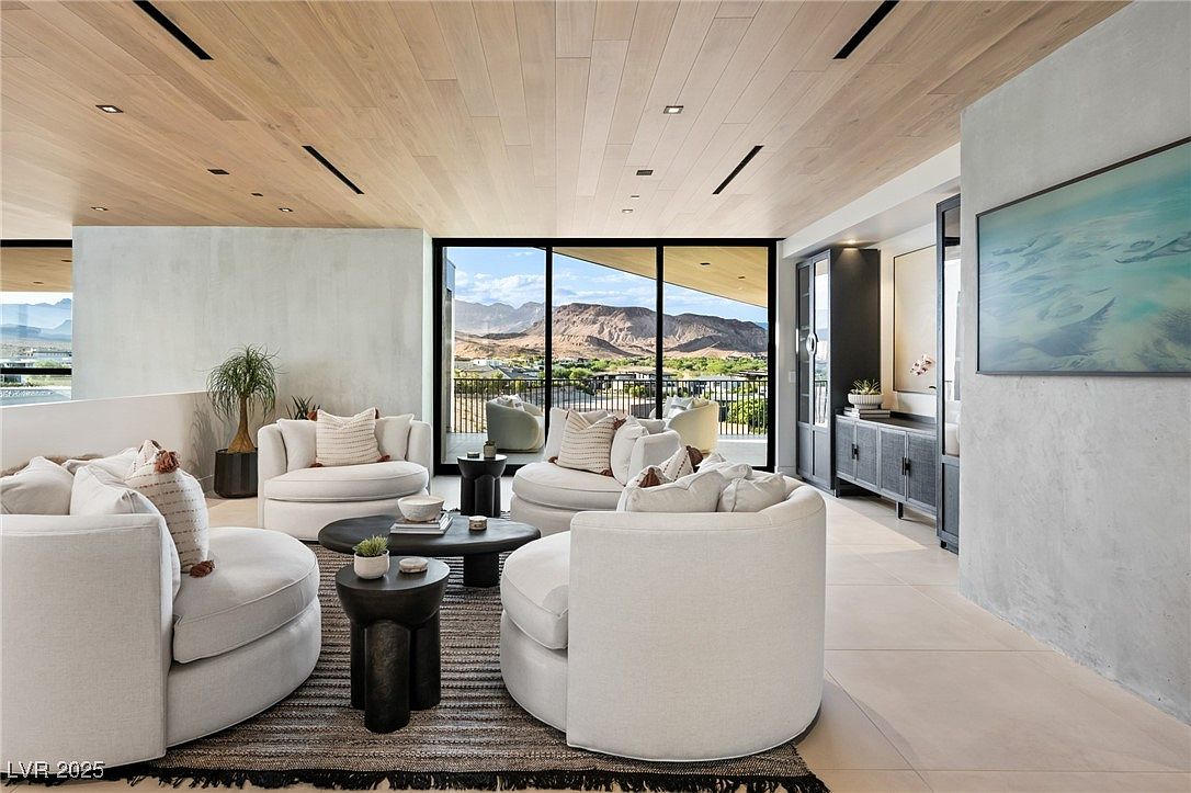 This is a bright and airy living room featuring modern design elements. The room is furnished with several round, white upholstered chairs arranged around a dark, round coffee table and smaller side tables, all set on a striped rug. Large windows offer a stunning view of the mountains, enhancing the room's appeal and creating a serene atmosphere.