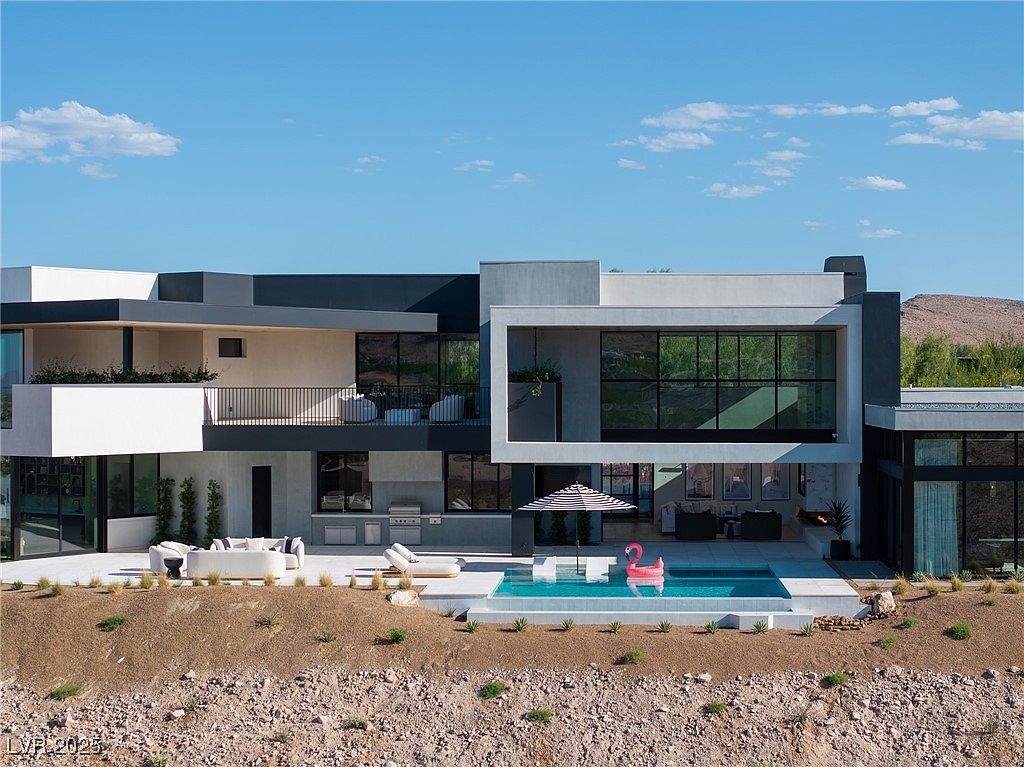 This image showcases the rear exterior of a modern luxury home, emphasizing its outdoor living space. A sleek pool with a flamingo floaty is the focal point, complemented by a patio area with lounge furniture and an outdoor kitchen. The architectural design features clean lines, large windows, and a combination of light and dark gray tones, creating a sophisticated and inviting atmosphere.