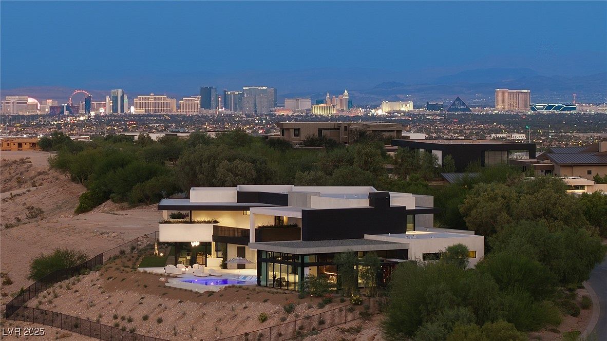 This image showcases the rear exterior of a modern luxury home with a pool and patio area. The architecture features clean lines, a mix of light and dark materials, and large windows offering views of the city skyline in the background. The landscaping is minimal, emphasizing the contemporary design and the elevated position of the property.