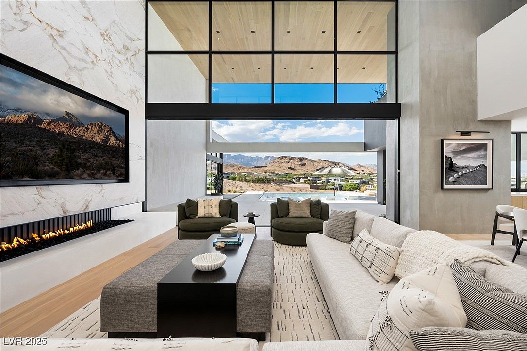 This is a luxurious living room featuring a modern fireplace with a large television above it, a comfortable sofa with decorative pillows, and two round armchairs. The room has floor-to-ceiling windows that offer a stunning view of the landscape outside, creating a seamless indoor-outdoor living experience. The overall design is contemporary and sophisticated, emphasizing natural light and open space.