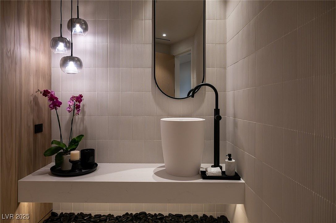 This is a stylish guest bathroom featuring a modern vessel sink, a sleek black faucet, and a minimalist mirror. The walls are adorned with textured, vertically-lined tiles, complemented by a wood-paneled section. Underneath the floating countertop, black pebbles add a unique decorative touch, and pendant lights provide soft illumination.