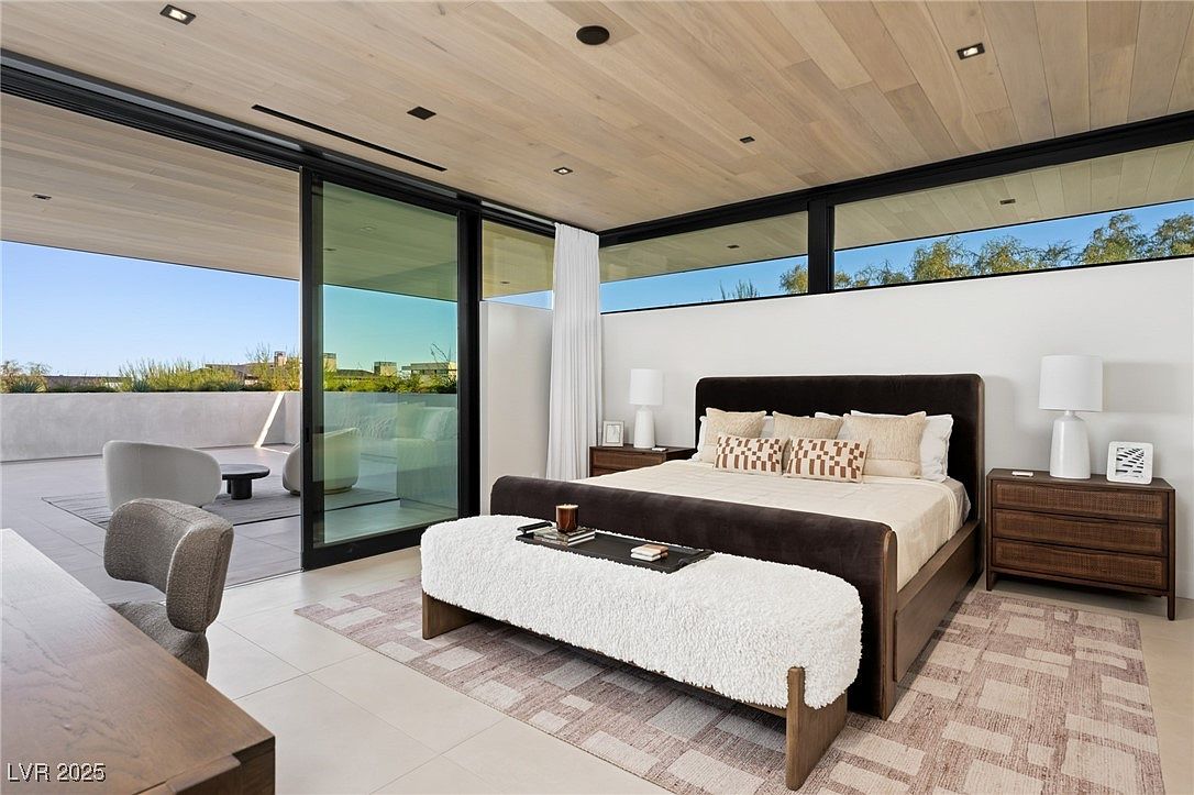 This is a primary bedroom featuring a modern design with a large bed, a bench at the foot of the bed, and nightstands on either side. Sliding glass doors open to an outdoor patio area, enhancing the sense of space and light. The room is decorated in neutral tones, creating a serene and luxurious atmosphere.