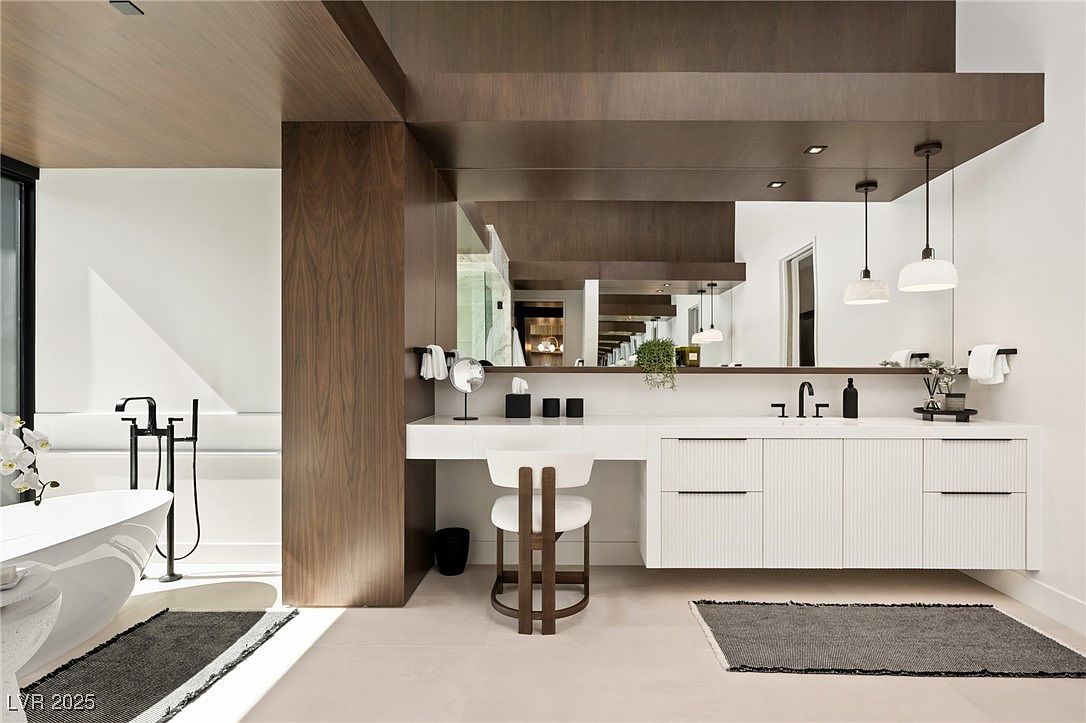 This is a modern primary bathroom featuring a sleek, floating vanity with white cabinetry and a dark wood accent wall. The space includes a freestanding bathtub with a black faucet, a large mirror reflecting the vanity area, and pendant lighting. The overall design is minimalist and luxurious, creating a spa-like atmosphere.