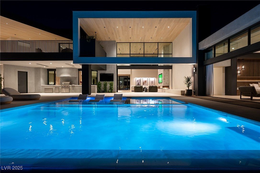 This image showcases a luxurious modern home with a stunning pool as the focal point. The pool features built-in lounge chairs and is illuminated with a vibrant blue light, creating an inviting atmosphere. The architecture is sleek and contemporary, with clean lines and large windows that offer views into the home's interior, including a living area and a second-story space with wood ceilings.
