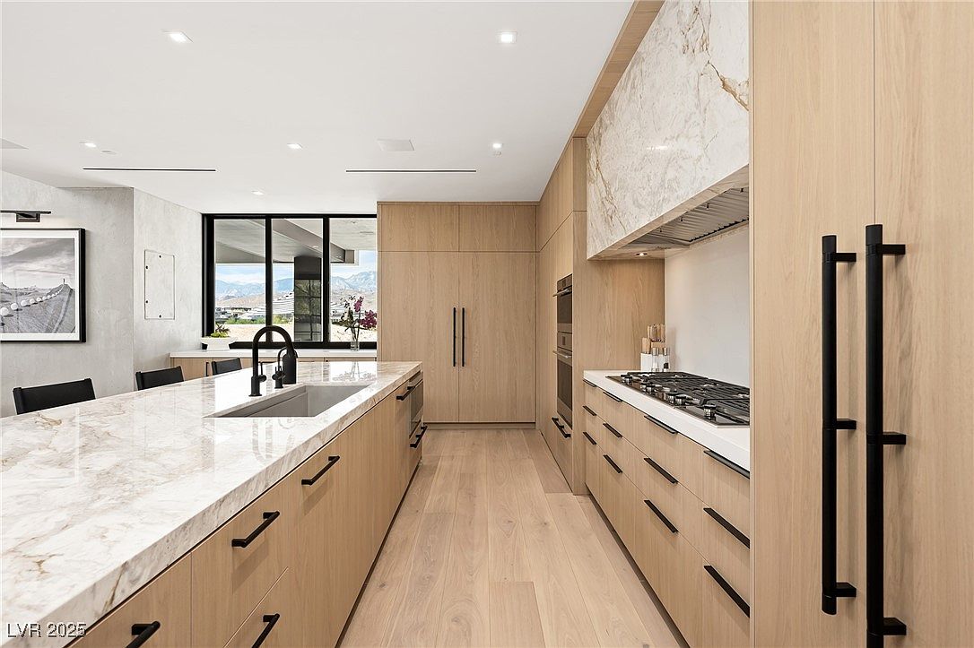 This is a modern kitchen with light wood cabinetry and black hardware. The countertops are a light marble with a large island featuring a sink and seating area. Stainless steel appliances are integrated into the cabinetry, and a large window provides natural light and a view of the outdoors. The perspective is from the entrance of the kitchen, showcasing the length of the island and the layout of the space.