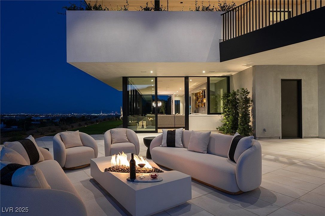 This image showcases a luxurious outdoor patio area with modern design elements. The space features comfortable seating around a contemporary fire pit, offering a cozy and inviting atmosphere. The patio is part of a modern home with large glass doors leading to the interior, and it overlooks a city view at dusk, enhancing the property's appeal for relaxation and entertainment.