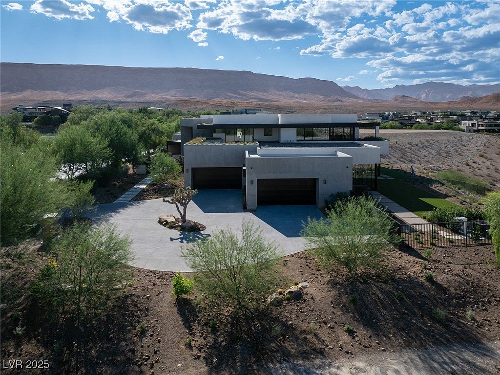 This is an exterior front view of a modern, two-story home with a flat roof and a neutral color palette. The house features a two-car garage and a circular driveway with desert landscaping. The backdrop includes mountains and a clear sky with scattered clouds, creating a serene and upscale ambiance.