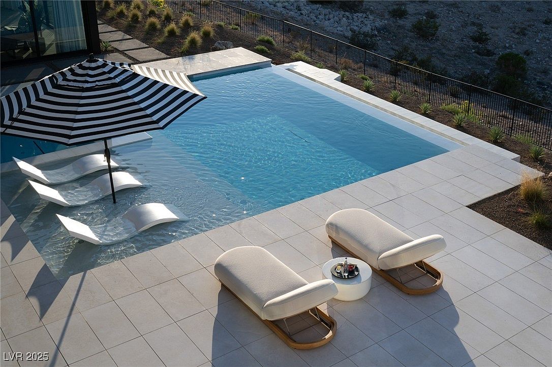 This image showcases a luxurious outdoor pool area, featuring a rectangular pool with clear blue water and integrated lounge chairs partially submerged. A striped umbrella provides shade, while two additional lounge chairs and a small table with drinks sit on the tiled patio. The setting is modern and upscale, suggesting a relaxing and sophisticated lifestyle.