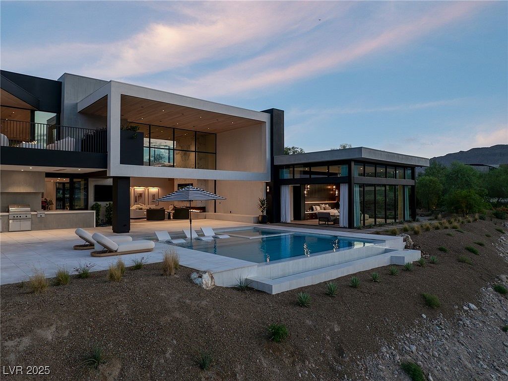 This image showcases the rear exterior of a modern luxury home, emphasizing the pool and outdoor living space. The pool is surrounded by a tiled patio with lounge chairs and an umbrella, while the architecture features clean lines, large windows, and a cantilevered second story. The landscaping is minimal, with natural terrain blending into the hillside, creating a serene and private setting.