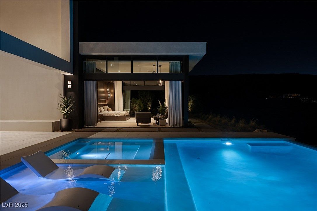 This image showcases a luxurious backyard pool and spa area at night. The pool features integrated lounge chairs and is illuminated with blue lighting, creating a serene ambiance. The modern architecture of the home is visible in the background, with large glass doors opening to a bedroom, seamlessly blending indoor and outdoor living.