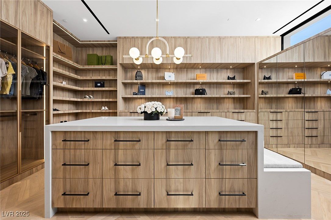 This is a luxurious walk-in closet featuring custom wood cabinetry and shelving, providing ample storage space for clothing and accessories. A central island with a white countertop offers additional storage and display space, while a modern chandelier illuminates the area. The overall design exudes sophistication and organization, perfect for a high-end real estate listing.