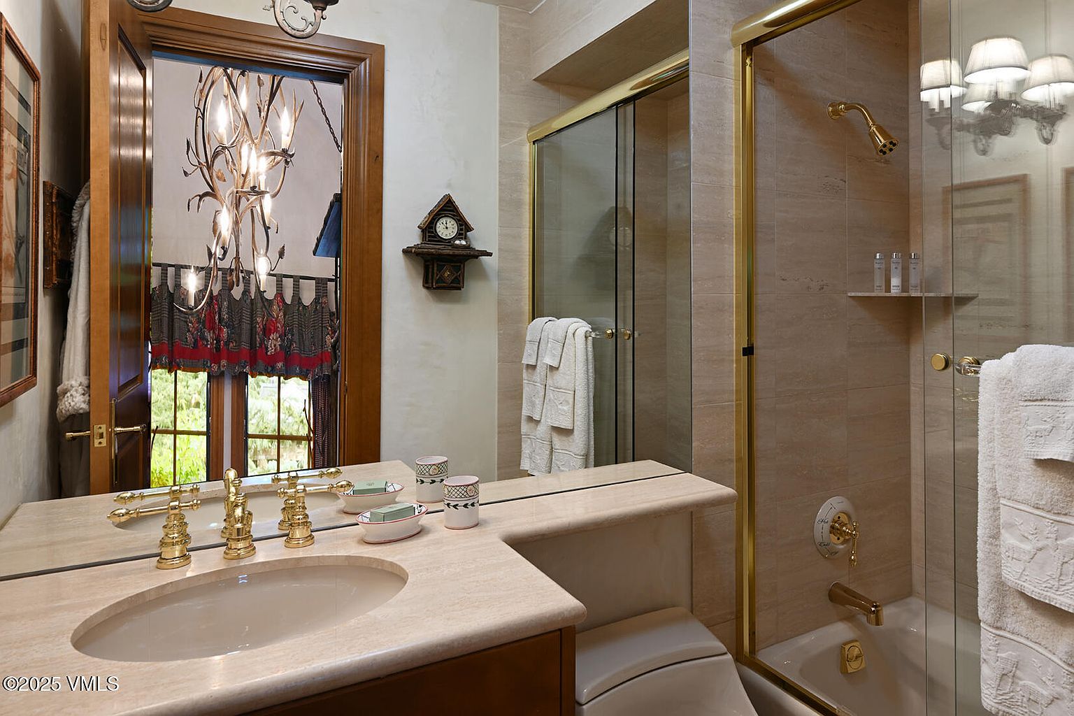 This is a bathroom featuring a beige color scheme with a vanity and sink, a mirror, and a shower/tub combination enclosed by glass doors with gold trim. A decorative clock hangs on the wall, and the overall style is traditional and well-maintained. The bathroom appears clean and functional, suitable for a guest or secondary bathroom.