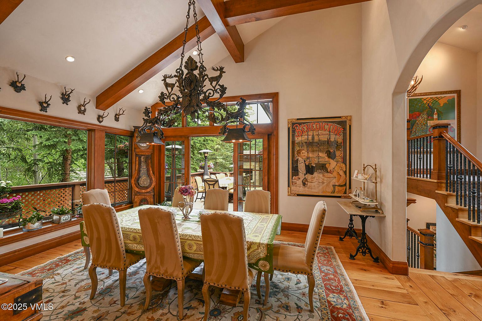 This is an interior shot of a dining room featuring a large wooden dining table with upholstered chairs. A decorative chandelier hangs above the table, and a vintage poster adorns the wall. The room has wooden beams on the ceiling and large windows offering a view of the outdoors, creating a warm and inviting atmosphere. The perspective is from the corner of the room, capturing the table, chandelier, and staircase.