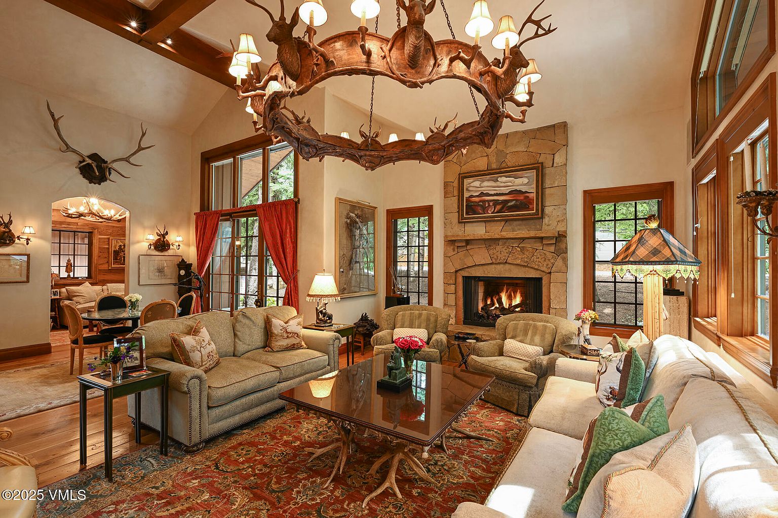This is a warm and inviting living room featuring a large stone fireplace, comfortable sofas, and a unique antler chandelier. The room is decorated in a rustic style with wood accents and a large area rug. The space is well-lit and appears to be a comfortable and relaxing area for entertaining or spending time with family.