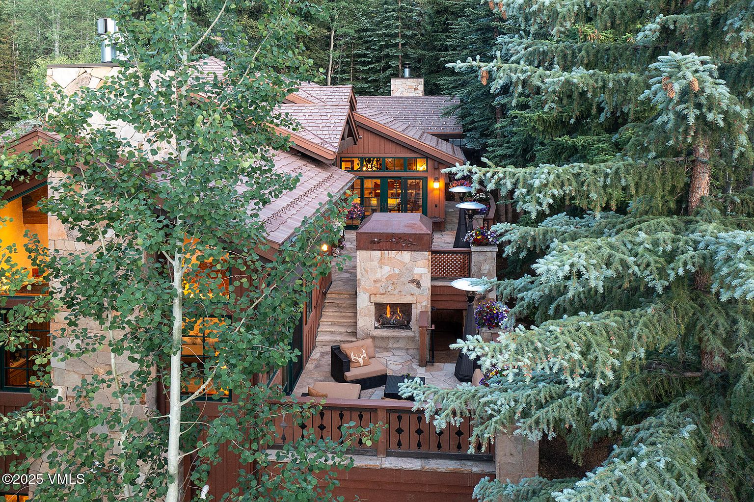 This image showcases the rear exterior of a luxurious mountain home, featuring a multi-level stone patio with a built-in fireplace and comfortable seating. The architecture blends seamlessly with the surrounding forest, with mature trees framing the view and adding to the sense of privacy and tranquility. Warm lighting emanates from the home's windows, creating an inviting ambiance.