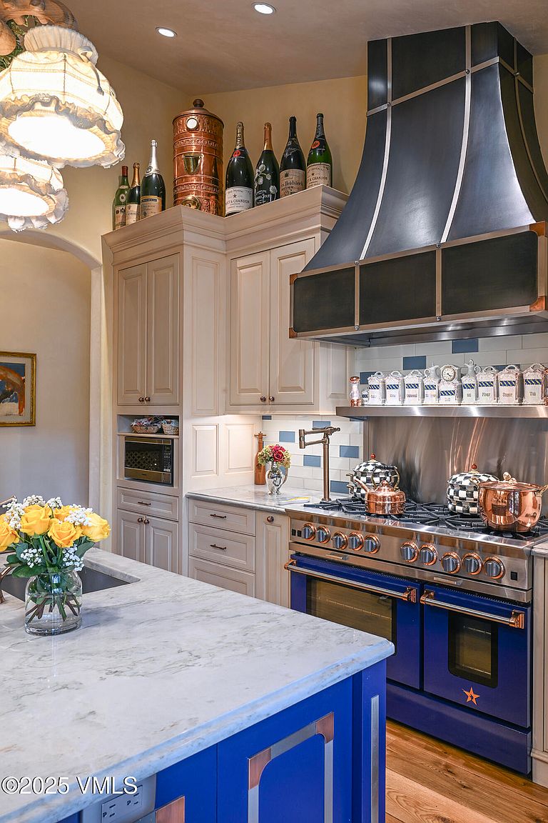 This is a well-appointed kitchen featuring custom cabinetry in a light cream color, complemented by a striking blue island and range. The countertops are a light marble, and the appliances are high-end, including a professional-grade range hood. Decorative items such as copper pots and wine bottles add a touch of elegance and personality to the space.