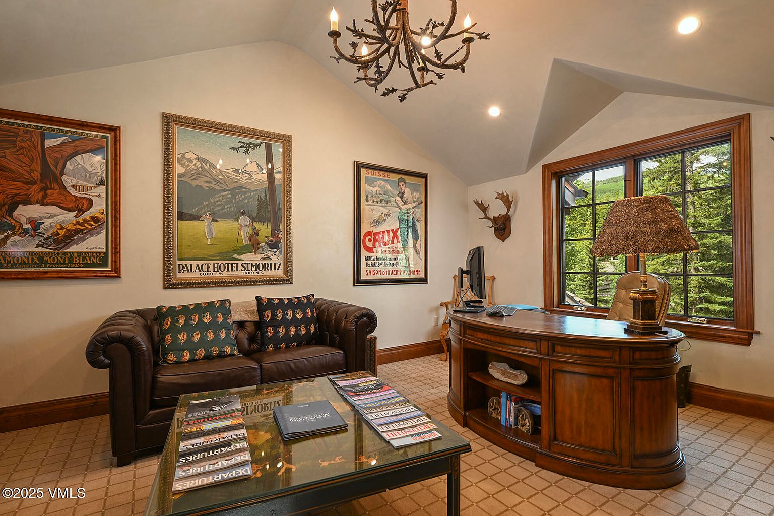 This is an interior shot of a study or office featuring a brown leather sofa with decorative pillows, a glass-topped coffee table with magazines, and a large wooden desk with a computer and lamp. The room is decorated with vintage travel posters and a rustic chandelier, creating a sophisticated and comfortable workspace with natural light from a window overlooking trees.