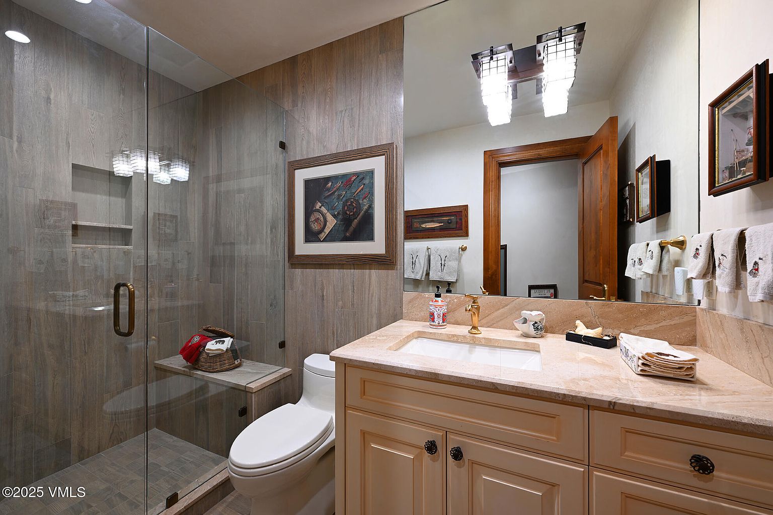 This is a well-appointed guest bathroom featuring a glass-enclosed shower with a built-in bench, a toilet, and a vanity with a light-colored countertop and cabinetry. The walls are covered in wood-look tile, and framed artwork adds a touch of personality. A large mirror above the vanity reflects the room, enhancing the sense of space.