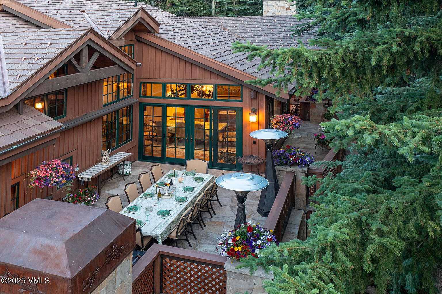 This image showcases a beautifully arranged outdoor patio area, perfect for entertaining. A long dining table is set for a meal, surrounded by comfortable chairs. The patio features stone flooring, elegant railings, and lush greenery, creating a serene and inviting atmosphere. The house has a rustic style with wood siding and green framed windows and doors.