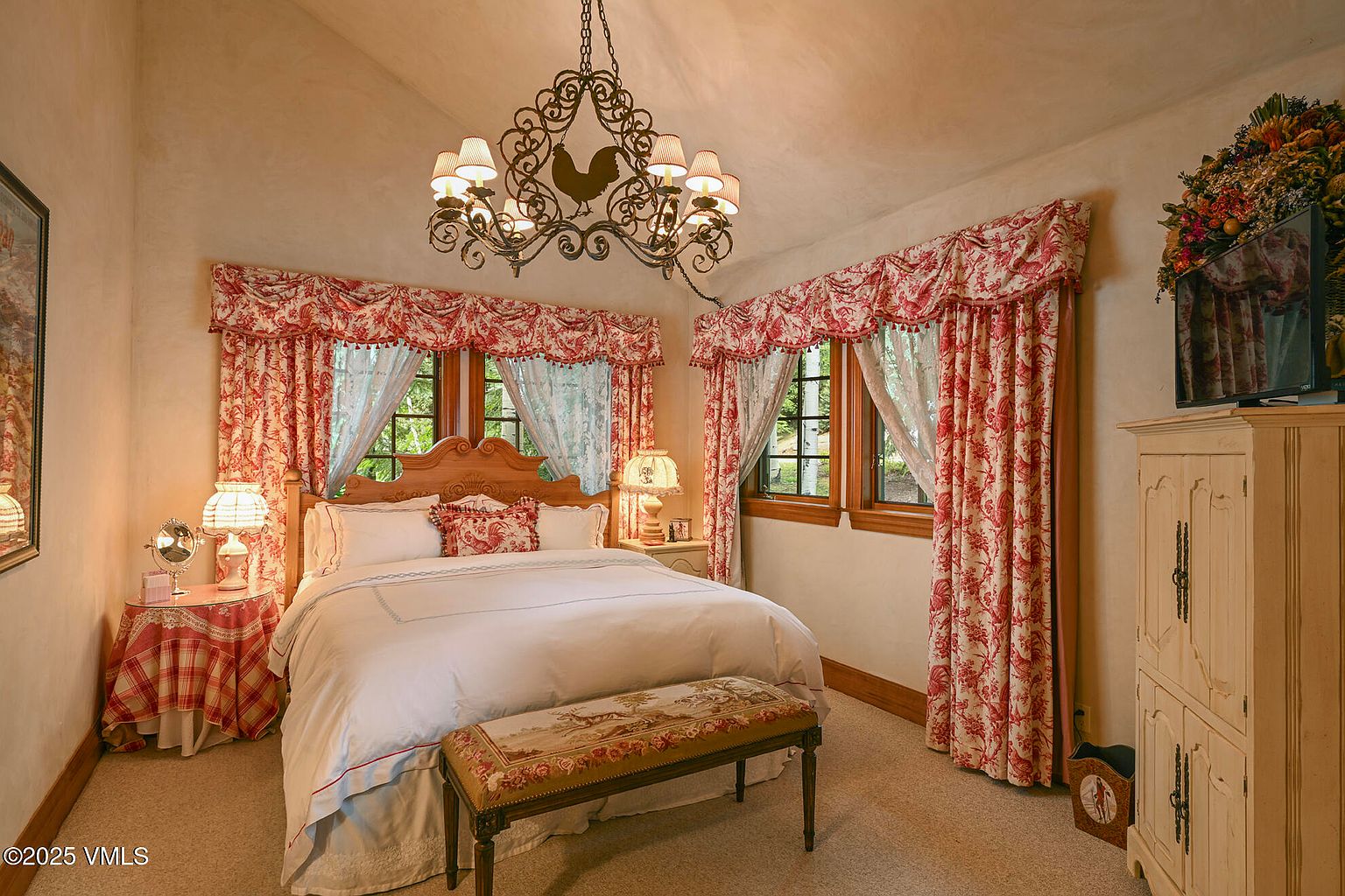This is a cozy primary bedroom featuring a large bed with white linens and decorative pillows, flanked by bedside tables with lamps. The room is adorned with red and white floral patterned curtains and valances, and a unique chandelier hangs from the ceiling. A decorative bench sits at the foot of the bed, and a wooden cabinet is visible in the corner, creating a warm and inviting atmosphere.