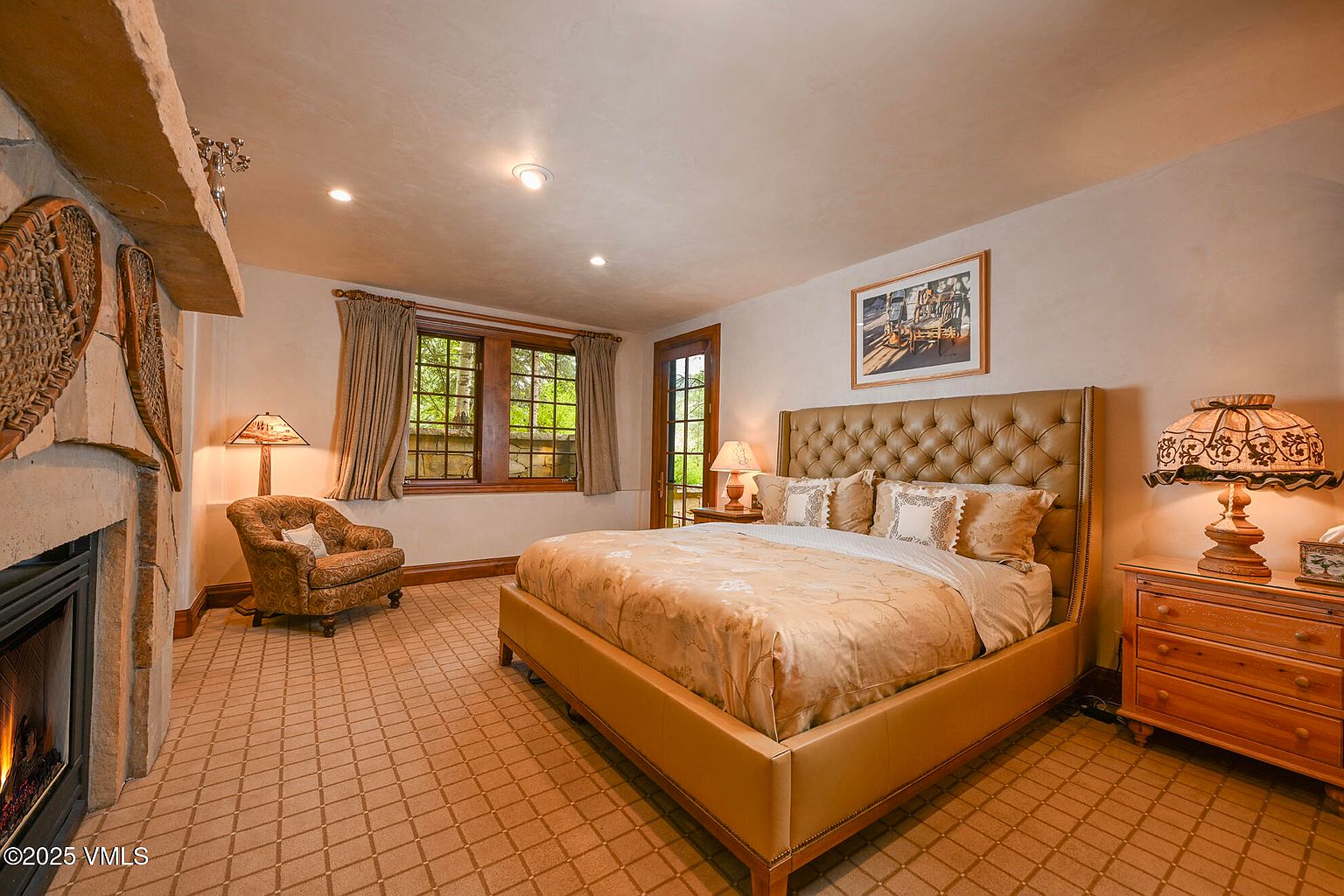 This is a primary bedroom featuring a large, tufted headboard bed with neutral-toned bedding. The room has a cozy and traditional feel, with a fireplace, a patterned armchair, and a wooden dresser with a decorative lamp. The walls are painted in a warm, neutral color, and the carpet has a geometric pattern.