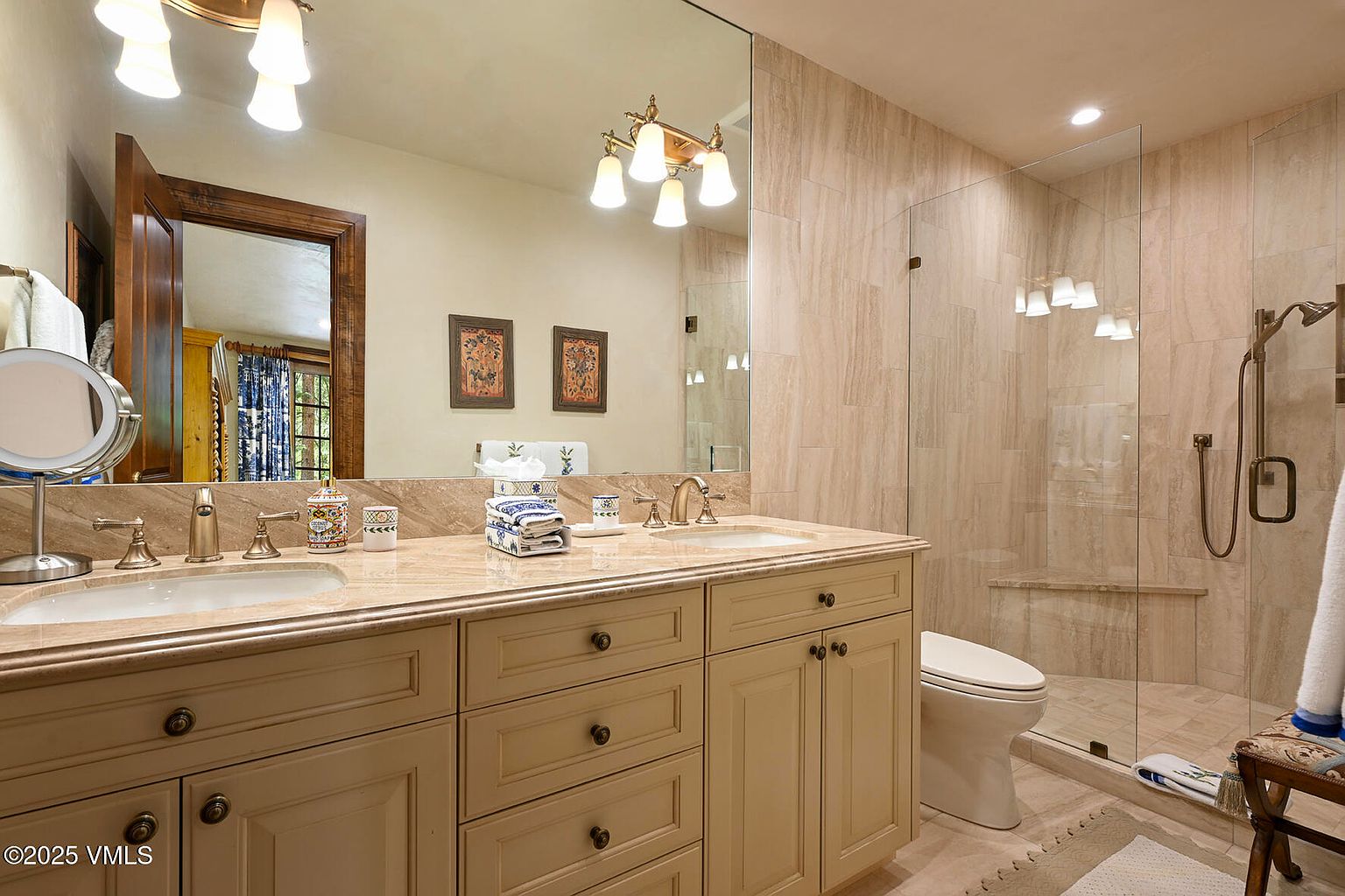 This is a well-lit primary bathroom featuring a double vanity with a light-colored countertop and cabinetry. A large mirror spans the length of the vanity, reflecting the lighting fixtures and artwork. The bathroom includes a glass-enclosed shower with tiled walls, and a toilet is visible to the side. The overall impression is clean, elegant, and functional.