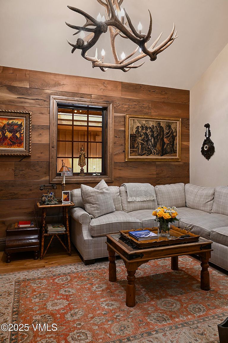This is an interior shot of a living room featuring wood paneled walls, a plaid sectional sofa, and a rustic coffee table. Artwork adorns the walls, and a unique antler chandelier hangs from the ceiling. The room exudes a warm, inviting, and sophisticated lodge-like atmosphere.
